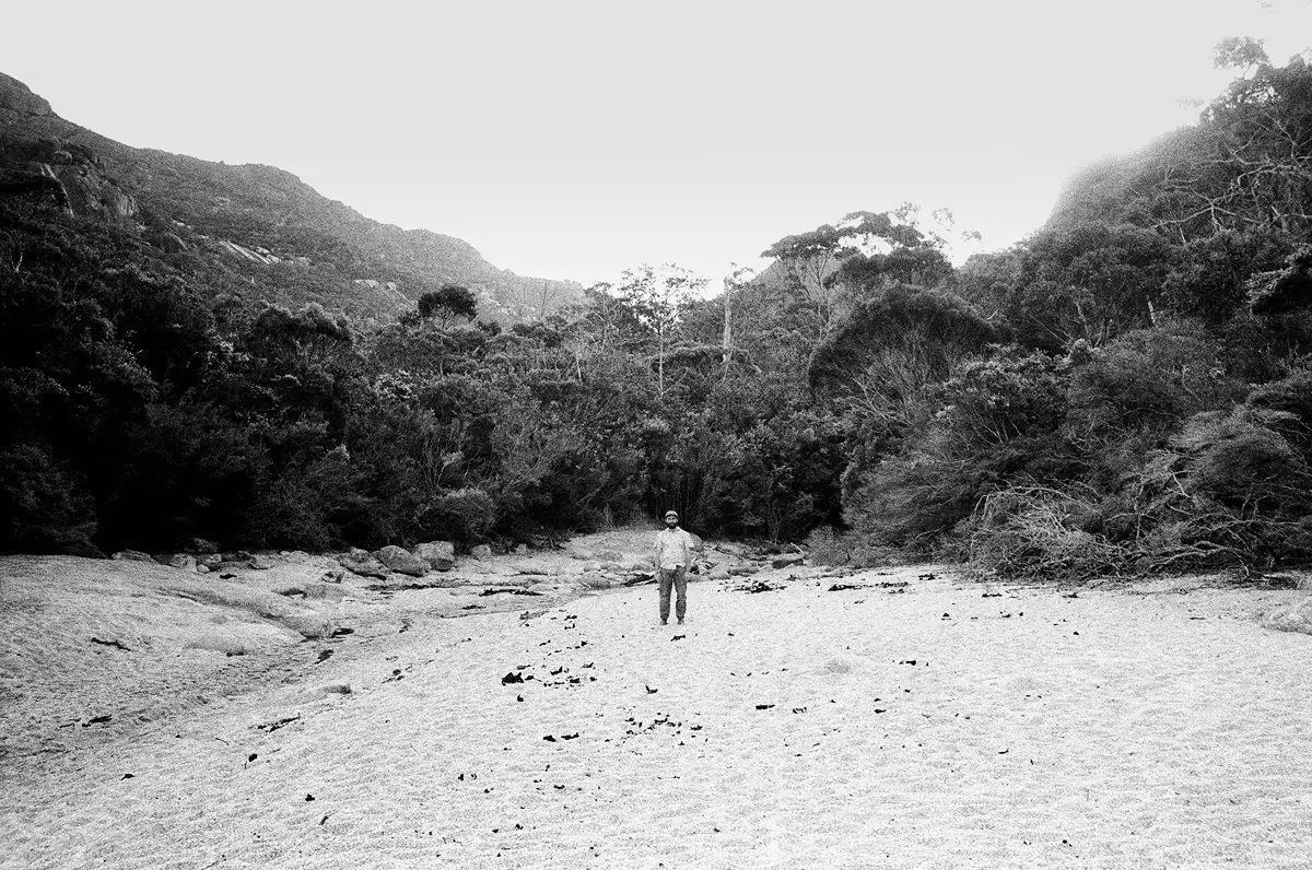 Lone figure standing on sandy riverbed surrounded by forested hills
