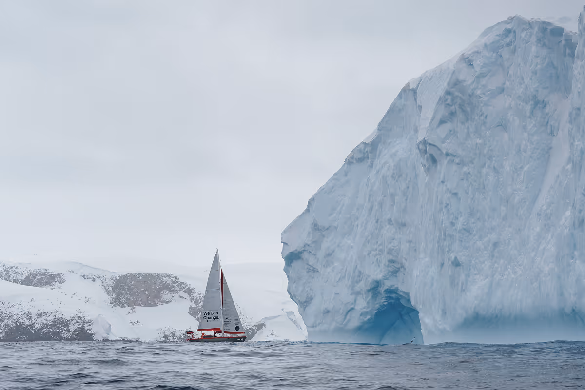 Sailboat passing towering blue iceberg with snow-covered mountains behind