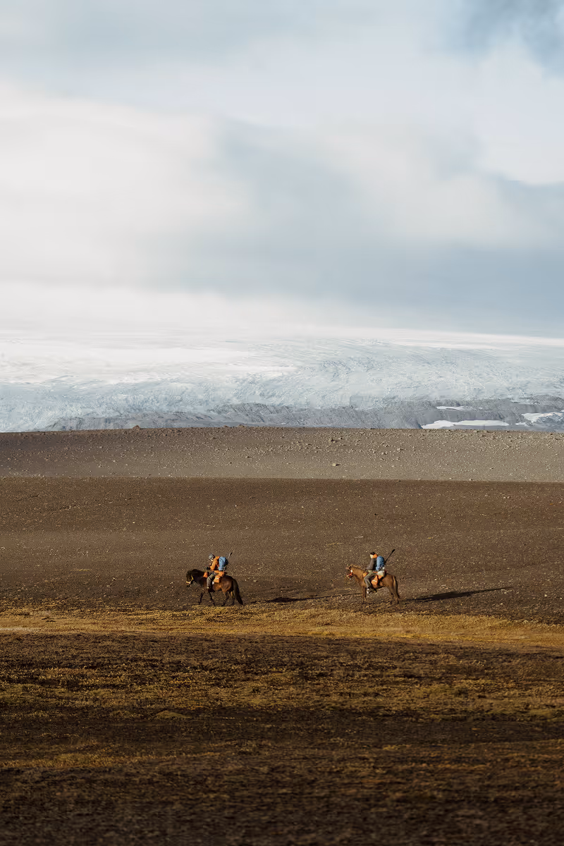 Two horseback riders crossing barren landscape with glacier in background