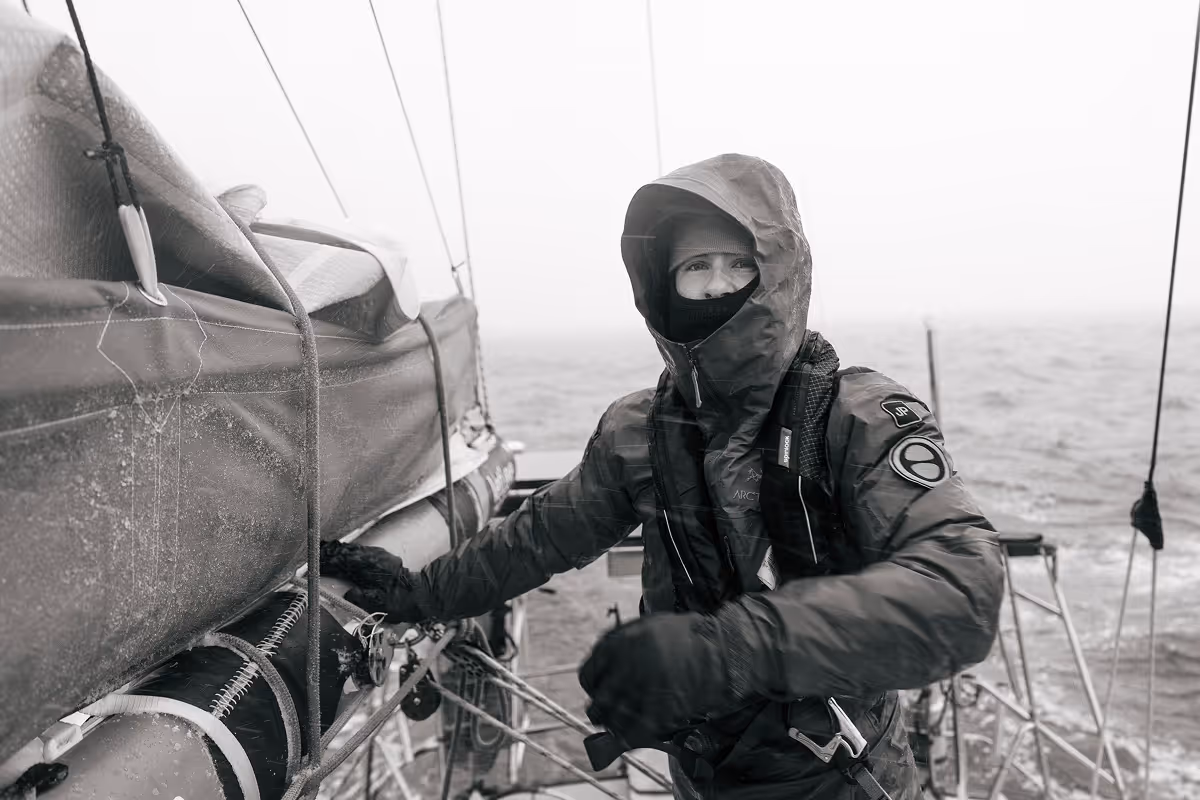 Person in heavy rain gear operating ropes on a sailboat in rough sea conditions.
