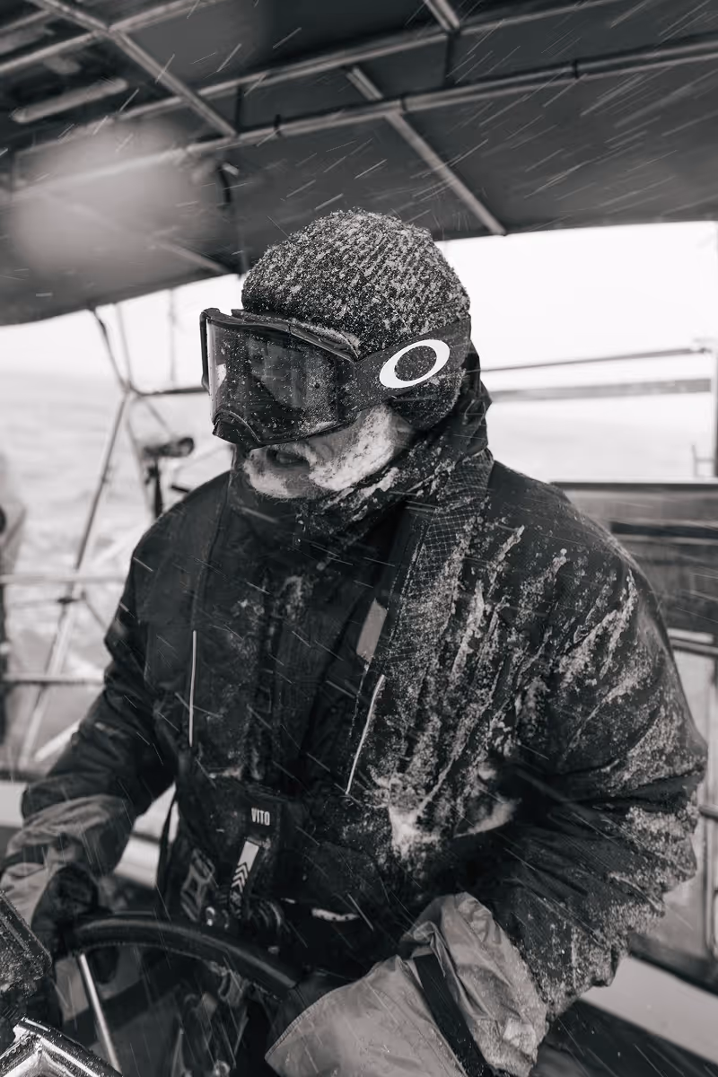 Person wearing winter gear and goggles steering a boat in snowy weather.