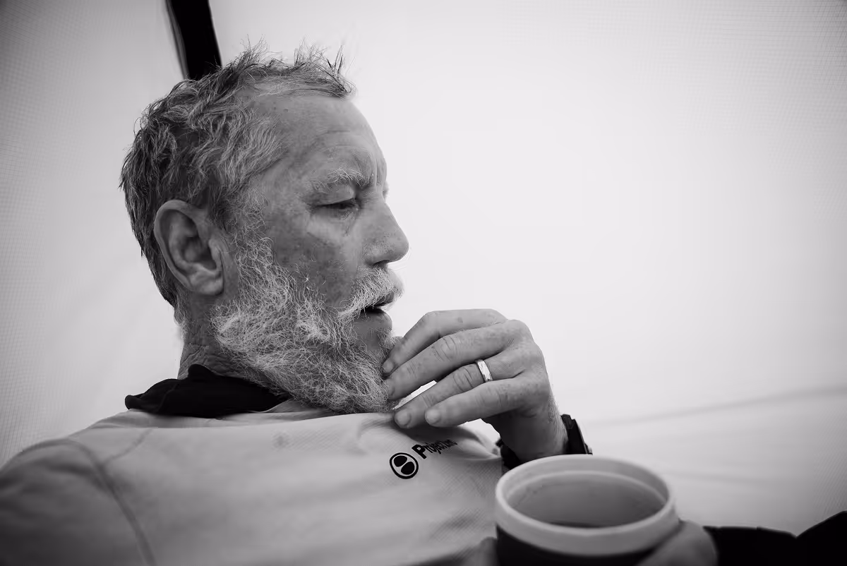 Black and white close-up of an older man with a beard holding a cup and resting his hand near his chin in thoughtful pose.