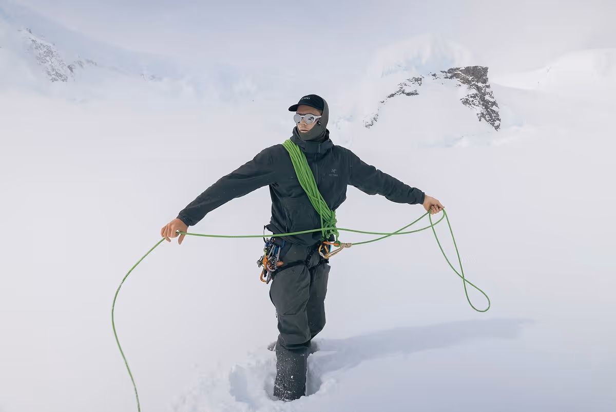Climber dressed in black winter gear holding and coiling a green climbing rope in a snowy mountainous landscape.