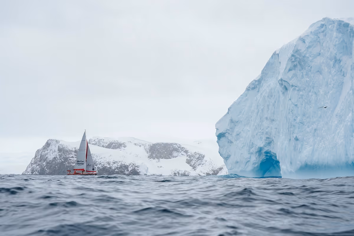 Red sailboat with 'We Can Change' text sailing near a large blue iceberg and snow-covered rocky shore under an overcast sky.