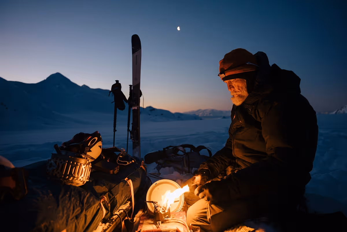 Man in winter gear lighting a small camp stove fire outdoors at dusk with skis and snowy mountains in the background.