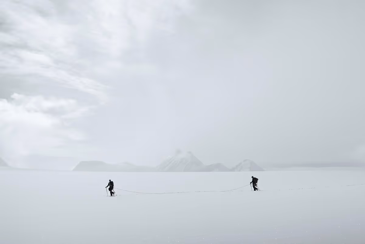 Two skiers connected by a rope traversing a vast snowy landscape with distant mountains under a cloudy sky.