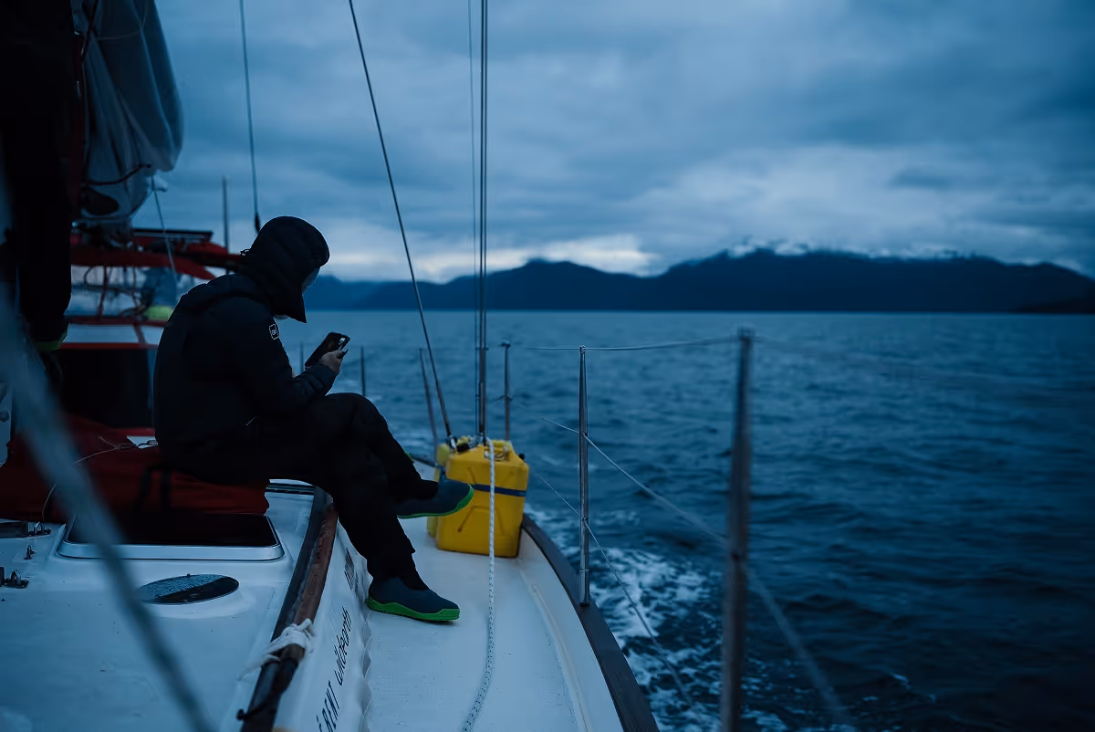 Person in black hooded jacket sitting on the edge of a sailboat looking at a phone during dusk on a calm sea with mountains in the background.