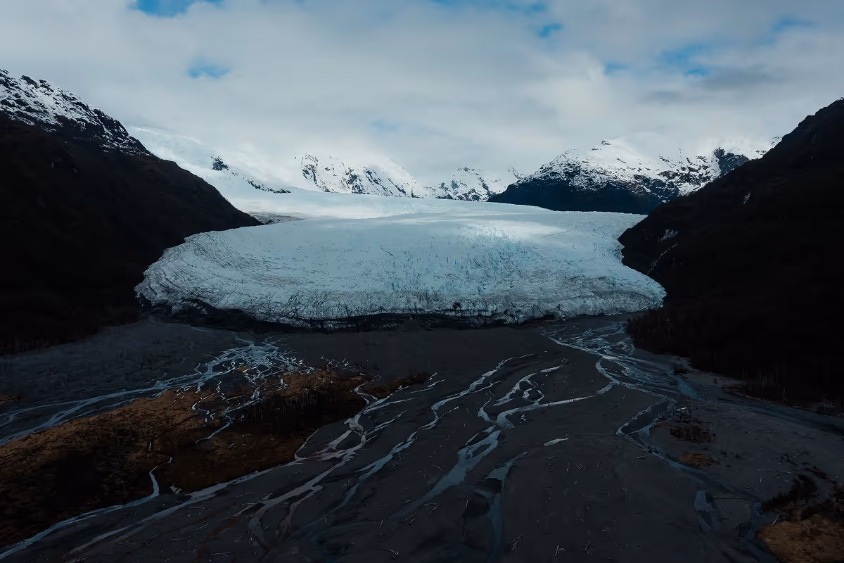 Large glacier extending between dark hills with snow-capped mountains under a cloudy sky.