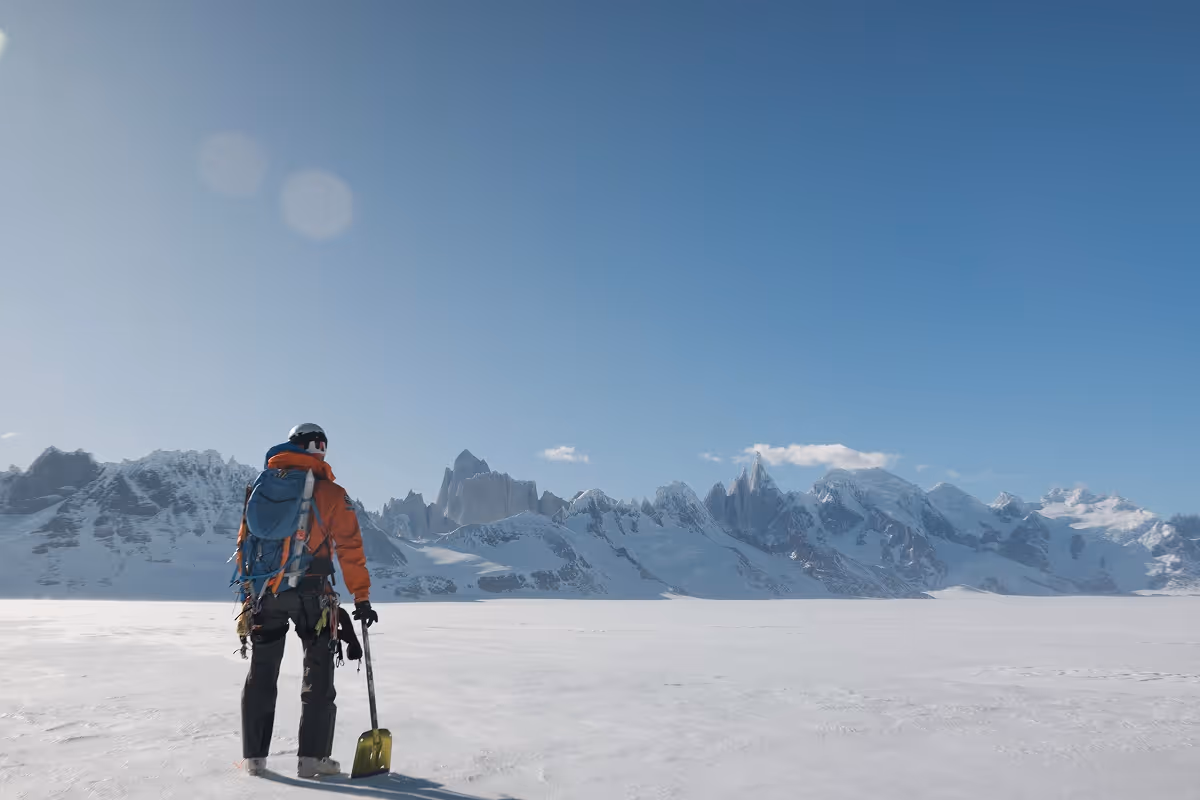 A lone mountaineer in orange jacket and helmet standing on a snowy plain holding a yellow shovel, facing distant snow-covered mountains under a clear blue sky.