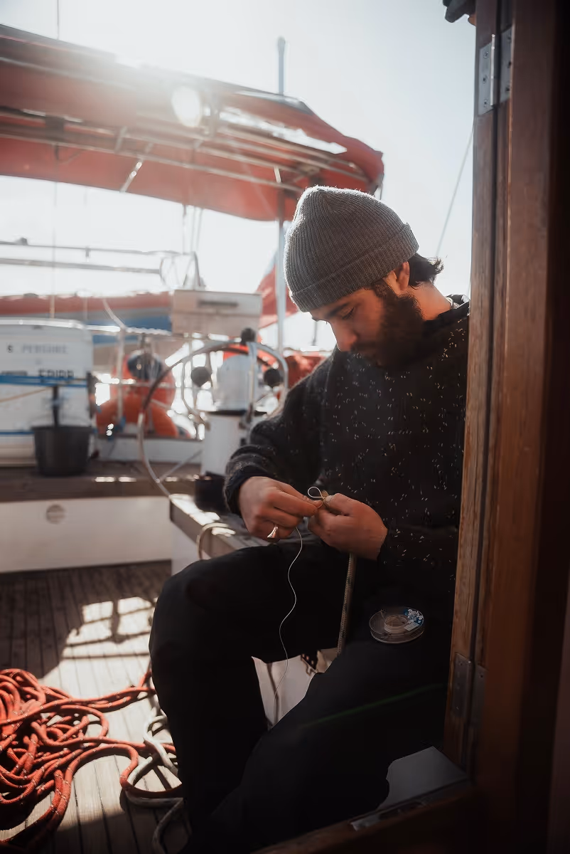 Bearded man wearing a gray beanie and dark sweater sitting on a boat deck, working with rope in his hands.