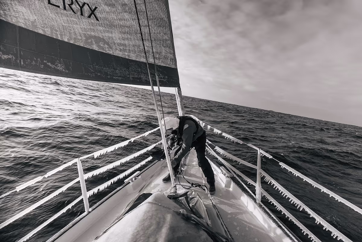 Person on a sailboat bow adjusting ropes with icicles hanging from the boat's railings and sail.