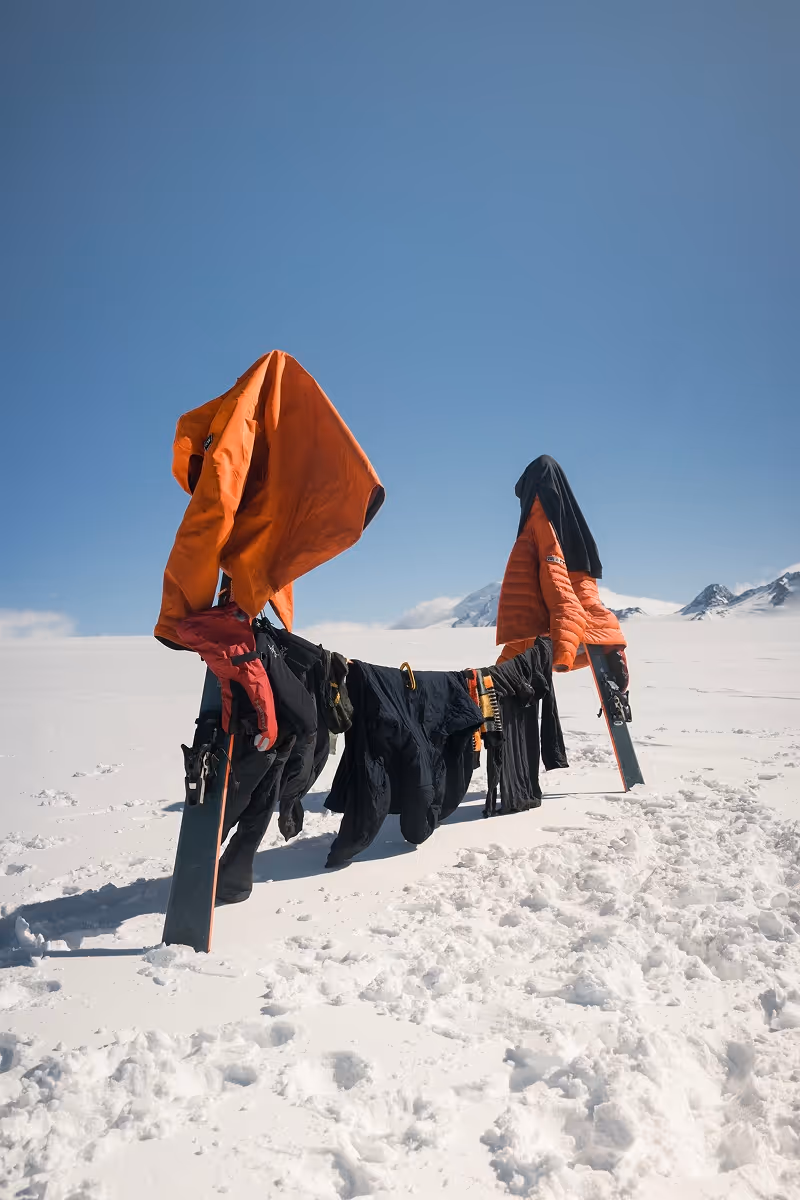Winter jackets and black clothing hanging on skis stuck in snow under clear blue sky in a snowy mountain landscape.