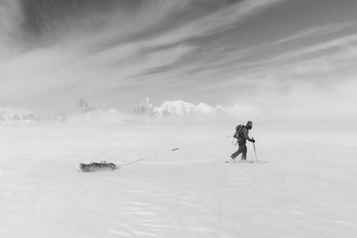 Skier pulling a sled across a snowy landscape with mountains and cloudy sky in the background.