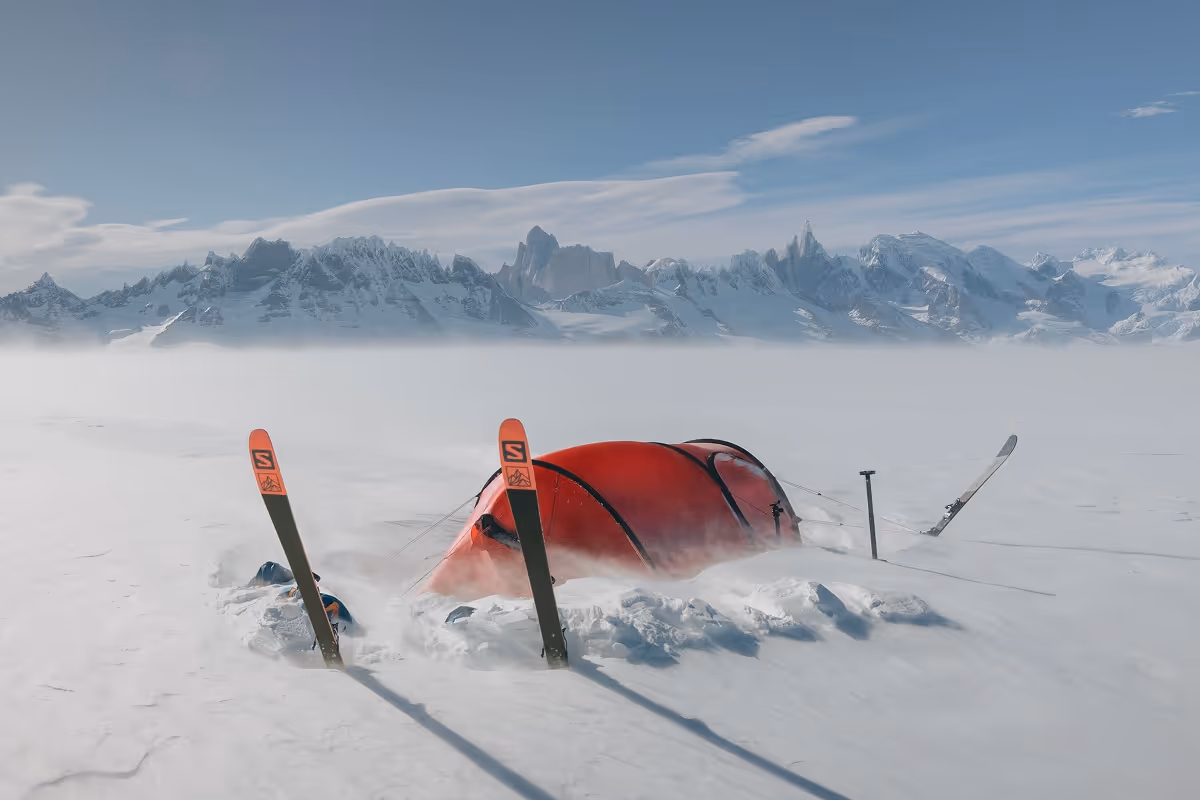 Red tent partially buried in snow on a snowy plain with skis planted upright around it and snow-covered mountains in the background under a clear blue sky.