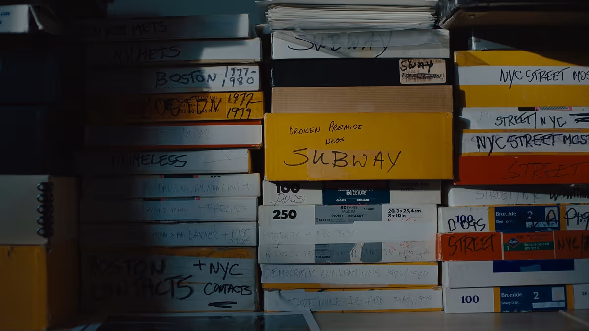 Stack of labeled cardboard boxes with handwritten titles including 'SUBWAY,' 'NYC STREET,' and dates, illuminated partly by light in a dim room.