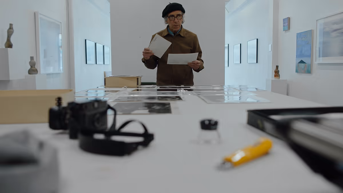 Man wearing glasses and a beret examines black-and-white photos laid out on a table in a bright gallery space.