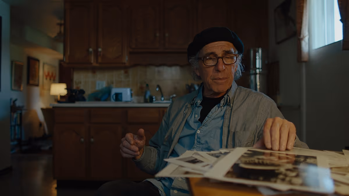 Elderly man wearing glasses and a black beret sitting at a table with vintage photographs, in a dimly lit kitchen.