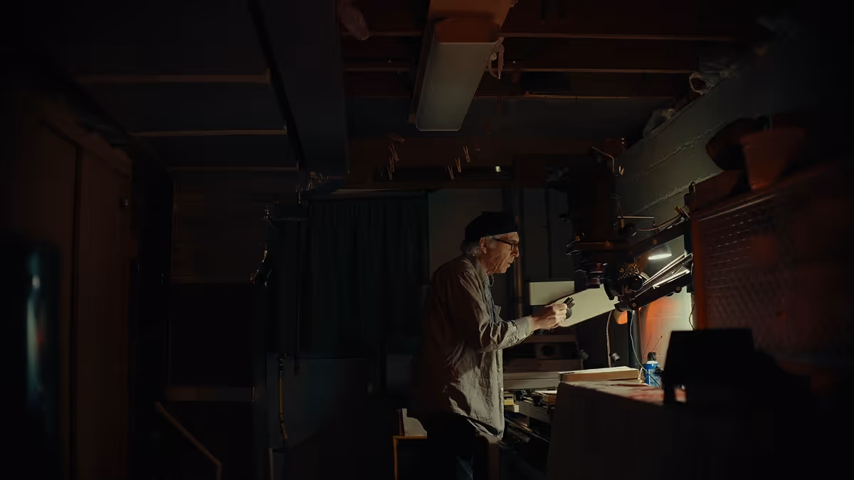 Man wearing glasses and a cap inspecting a photographic print under a light in a darkroom.