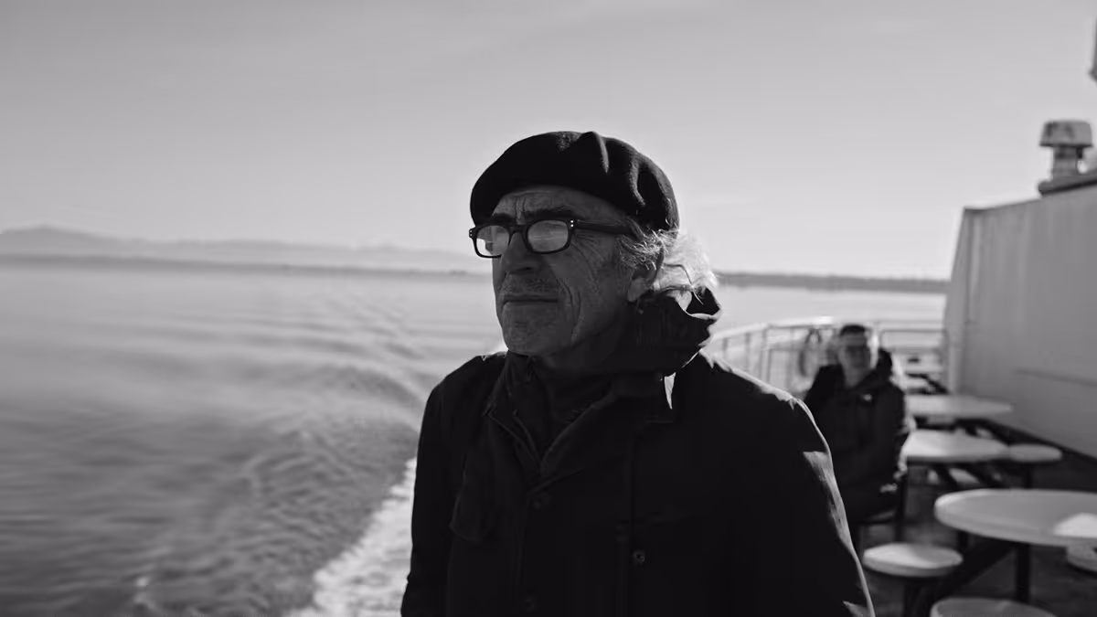 Black and white photo of an elderly man wearing a beret and glasses standing on a boat deck with water and distant mountains in the background.