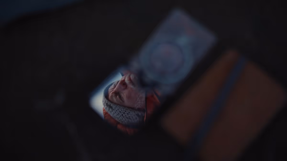 Reflection of a man wearing a knitted hat and an orange jacket in a rearview mirror.