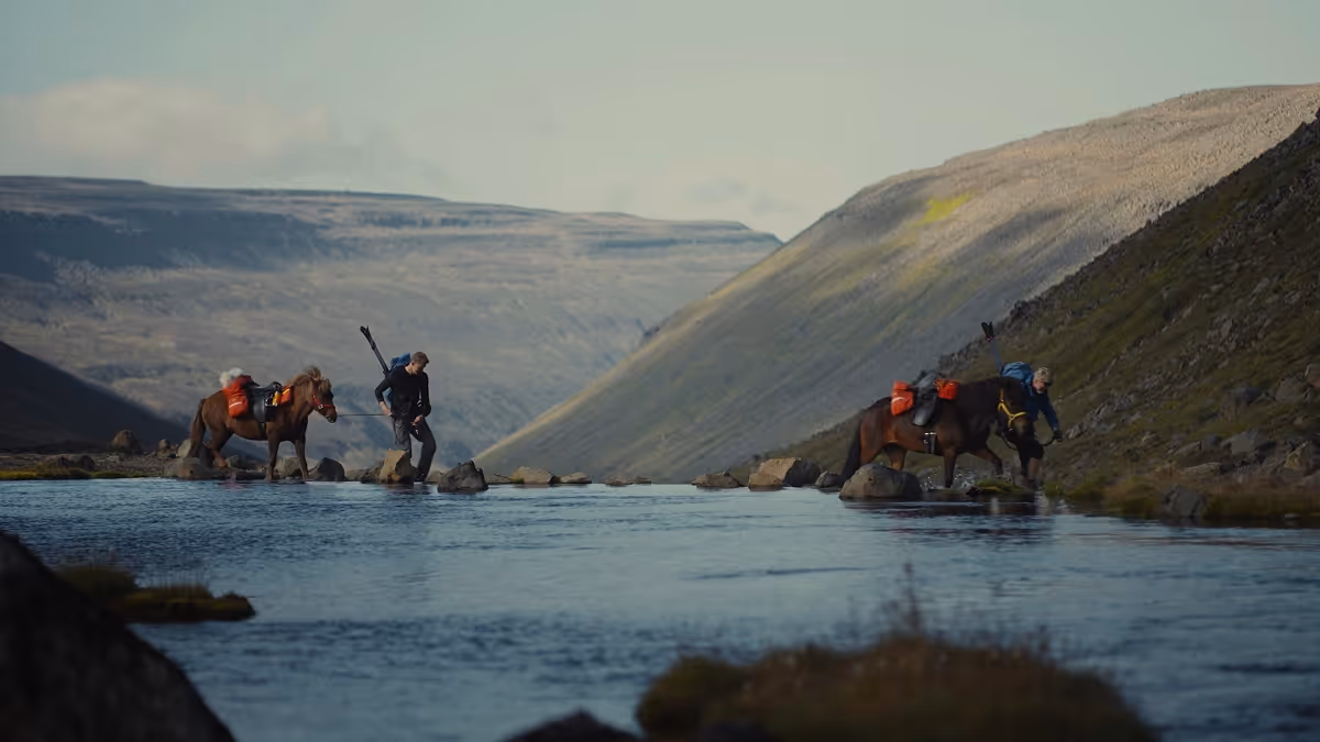 Two people with loaded horses crossing a shallow river in a mountainous landscape.