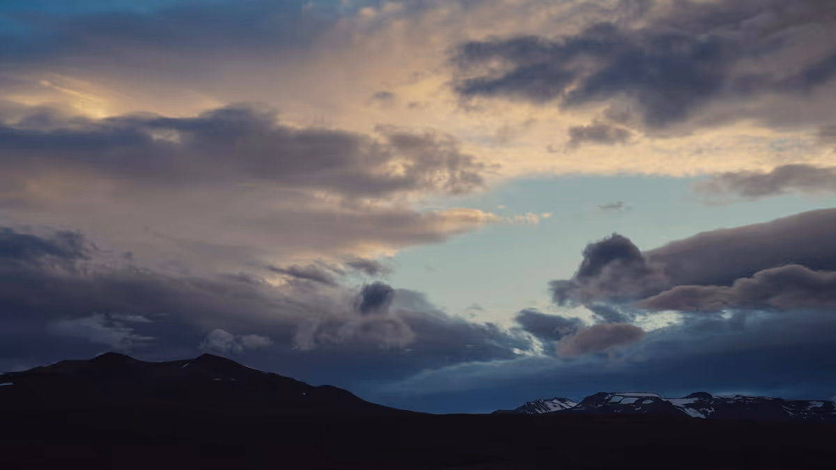 Dark mountain silhouettes under a dramatic cloudy sky with a small patch of blue light at sunset or sunrise.