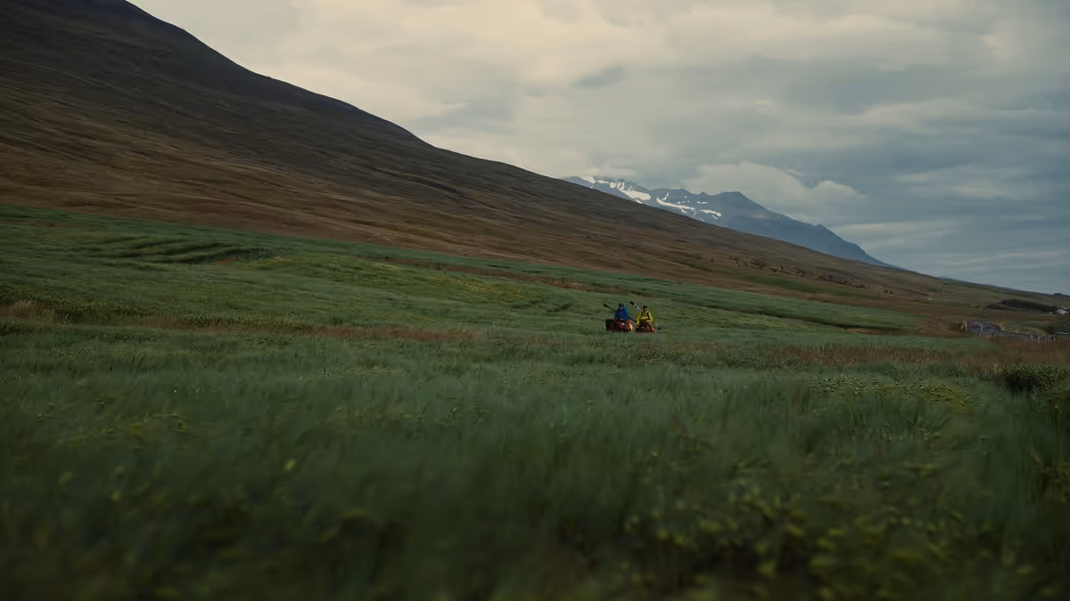 Two people kayaking on a river surrounded by green grass with mountains and a cloudy sky in the background.