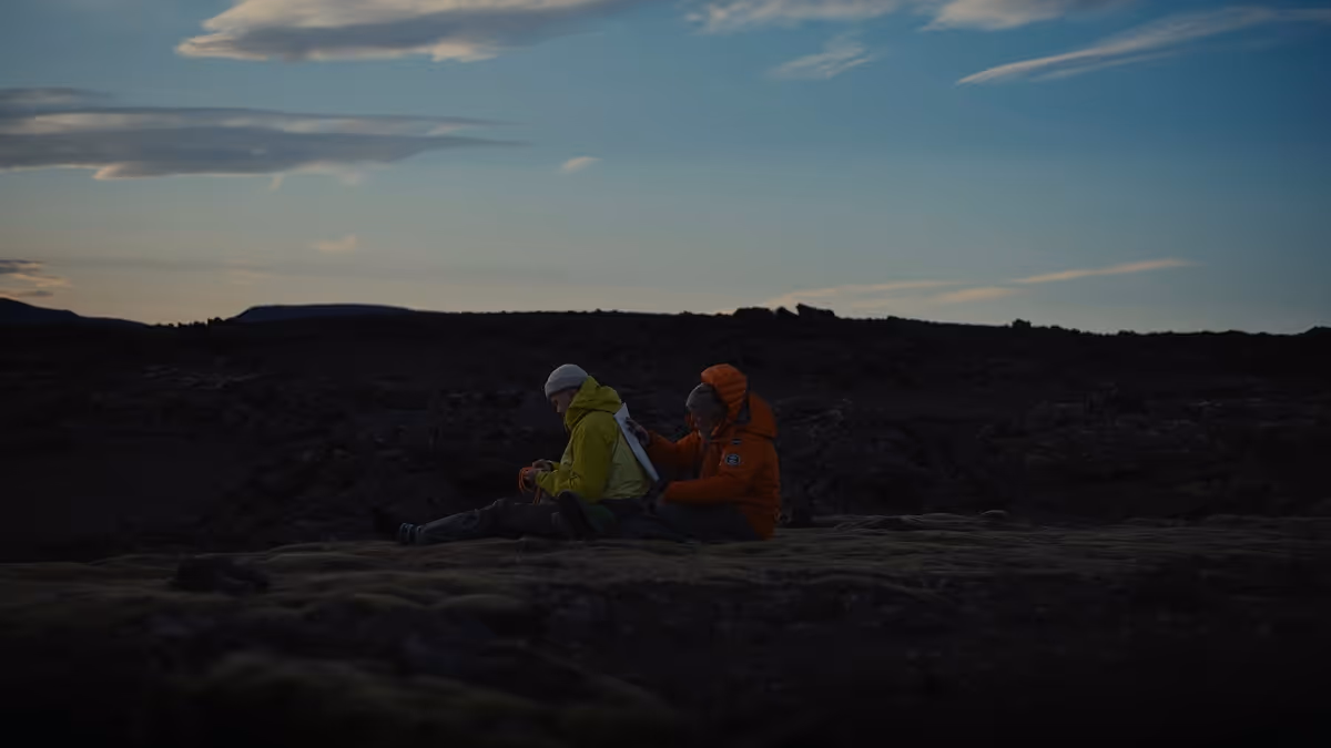 Two people sitting on rocky ground outdoors at dusk, one in a yellow jacket and the other in an orange jacket.
