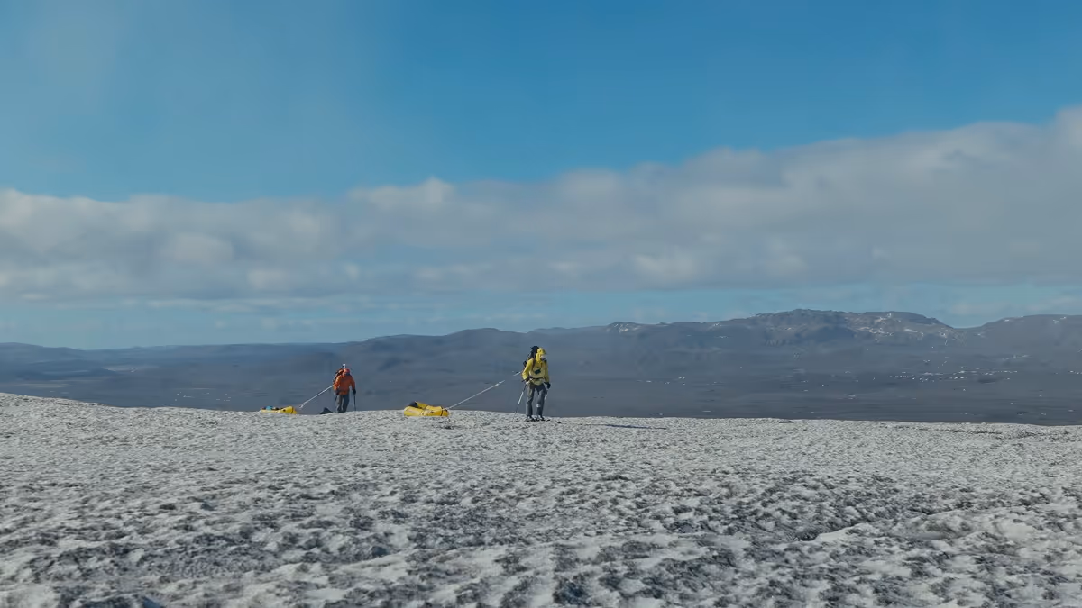 Two hikers walking across a snowy landscape, pulling yellow sleds with mountainous terrain in the background under a partly cloudy blue sky.