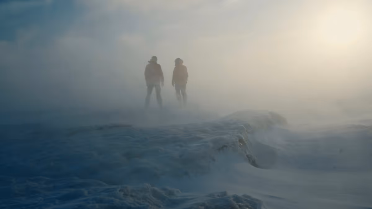 Two silhouetted people standing on a snowy landscape with mist and bright sunlight in the background.