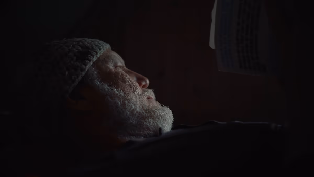 Side profile of an elderly man with a gray beard and knitted hat, lying down in dim light.