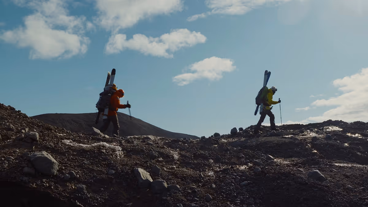 Two hikers carrying skis walk across a rocky, barren landscape under a partly cloudy blue sky.