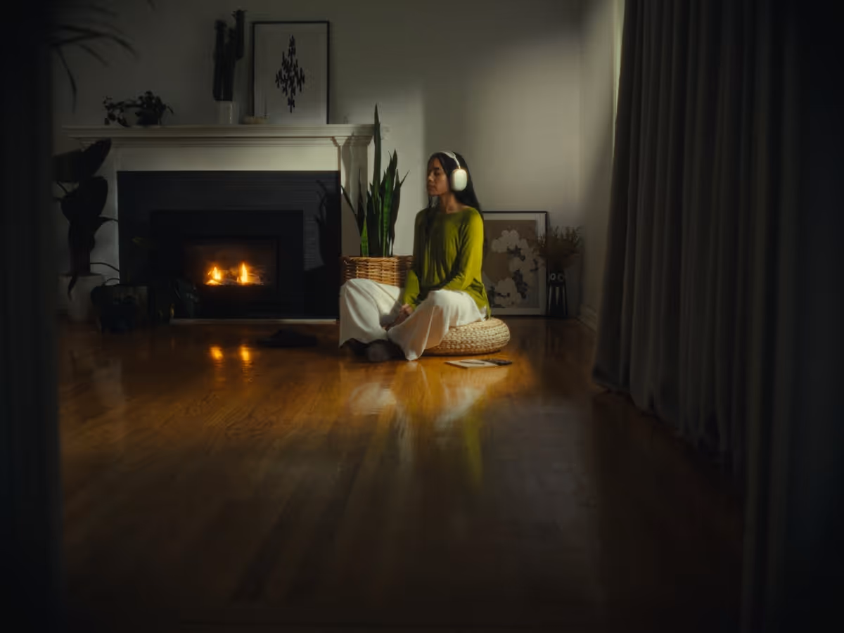 Woman sitting cross-legged on a cushion in a dimly lit room, wearing headphones near a lit fireplace with framed artwork and plants in the background.