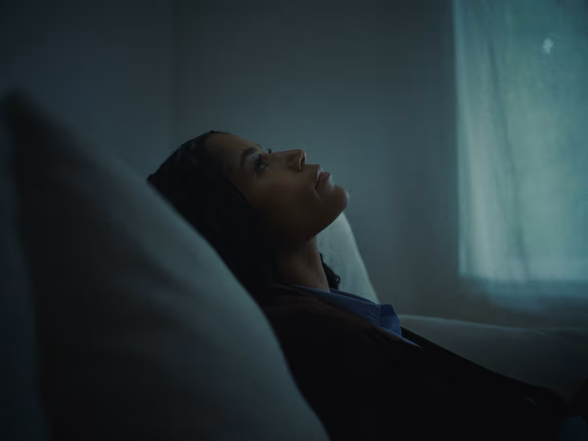 Woman reclining on a couch indoors, gazing thoughtfully towards a softly lit window.