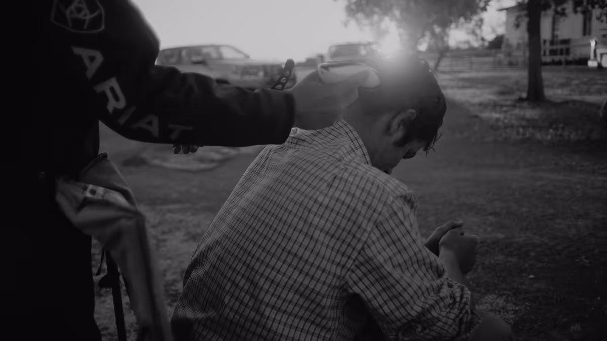 Black and white photo of a person holding a handgun to the head of a seated man outdoors with cars and trees in the background.