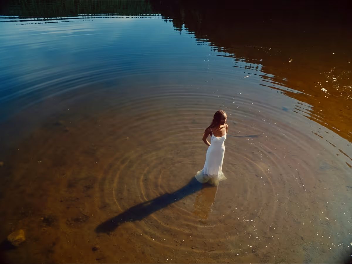 Woman in a white dress standing in shallow water with ripples and a long shadow at sunset.