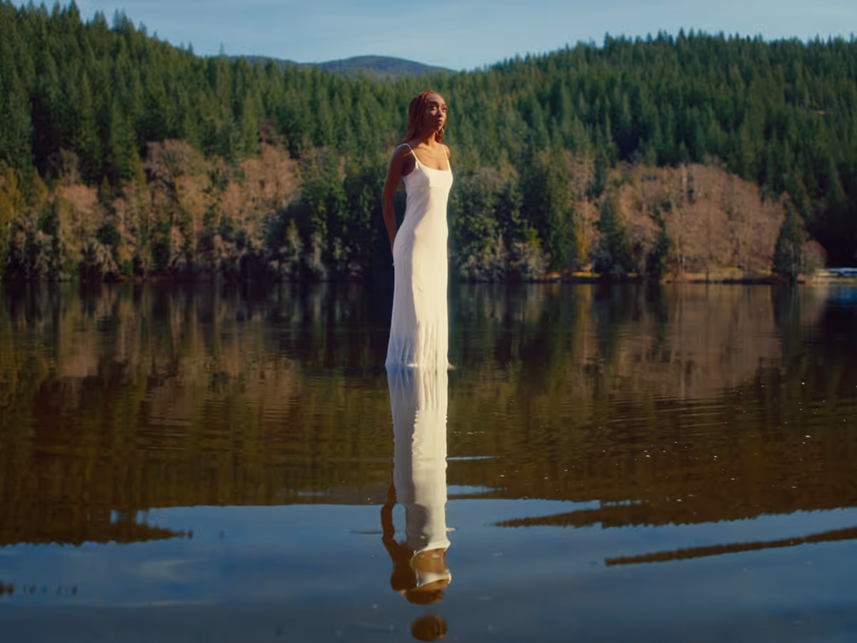 Woman in a white dress standing in calm water with forested hills in the background.