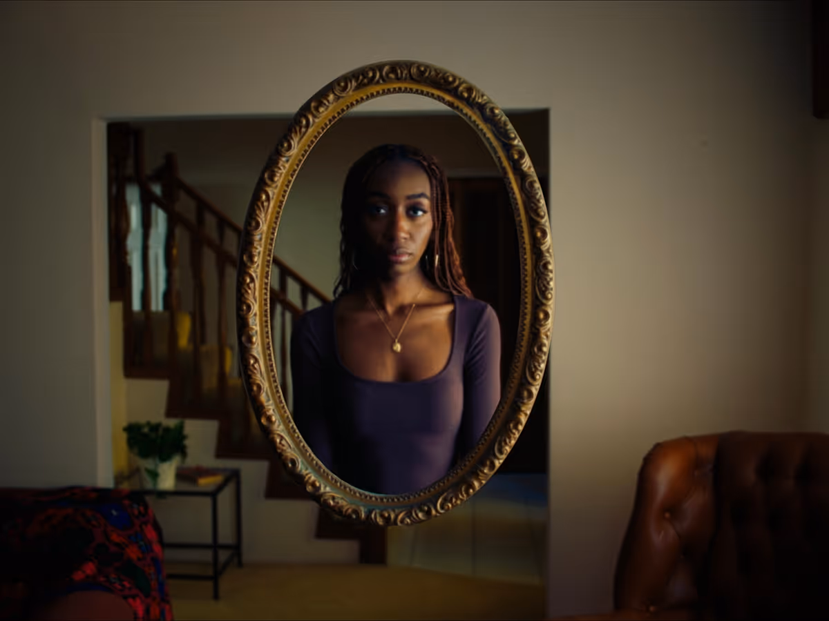 Woman with braided hair and purple top looking into an ornate oval mirror indoors.