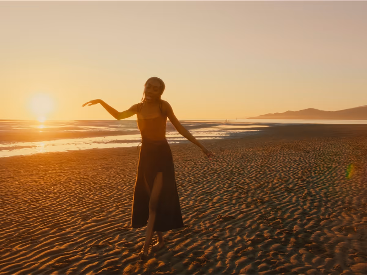 Person in dark dress walking barefoot on a rippled sandy beach at sunset with the sun low on the horizon.