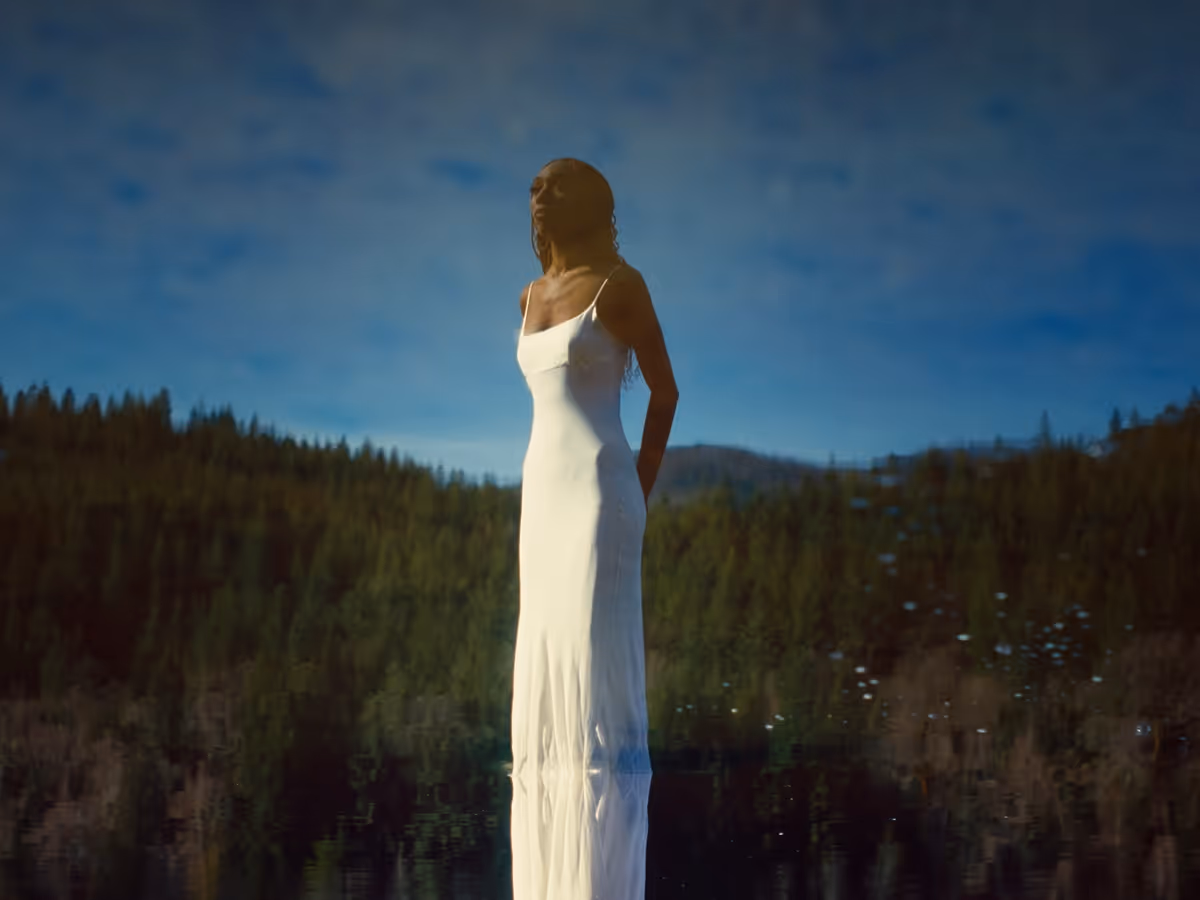 Woman in a long white dress standing outdoors with a forest and mountains in the background under a blue sky.