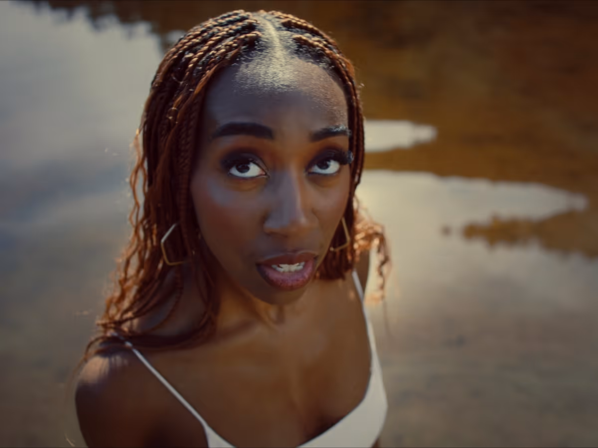 Close-up of a woman with braided hair and geometric earrings looking upwards, standing near reflective water.