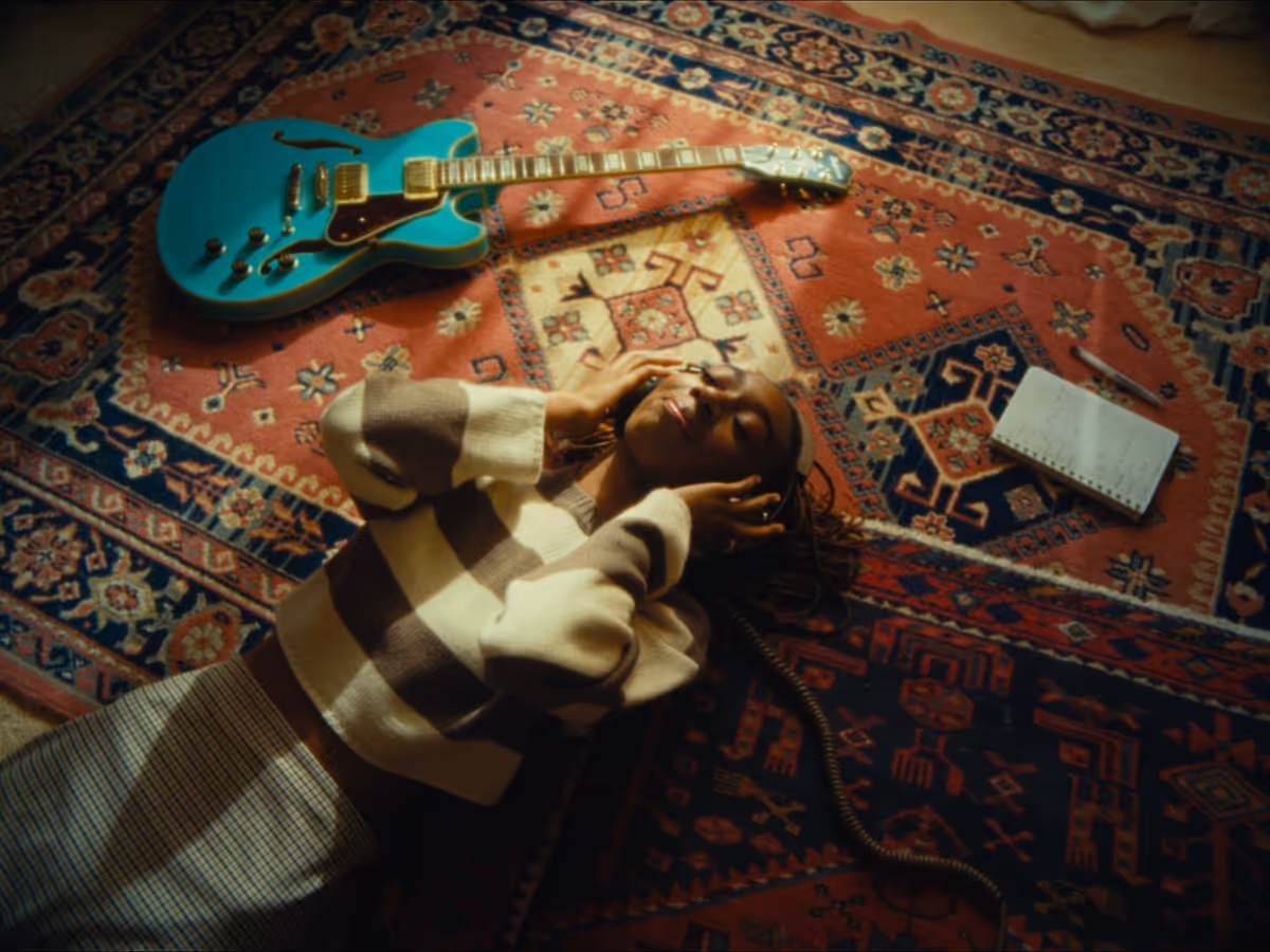 Person lying on a patterned rug with eyes closed, wearing headphones, next to a blue electric guitar and an open notebook with a pen.