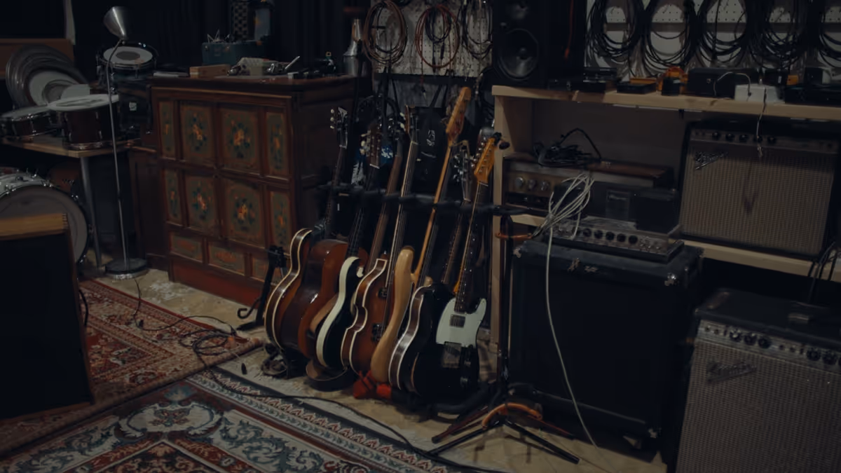 Six electric guitars on stands in a music studio with amplifiers, cables, and a drum set nearby.