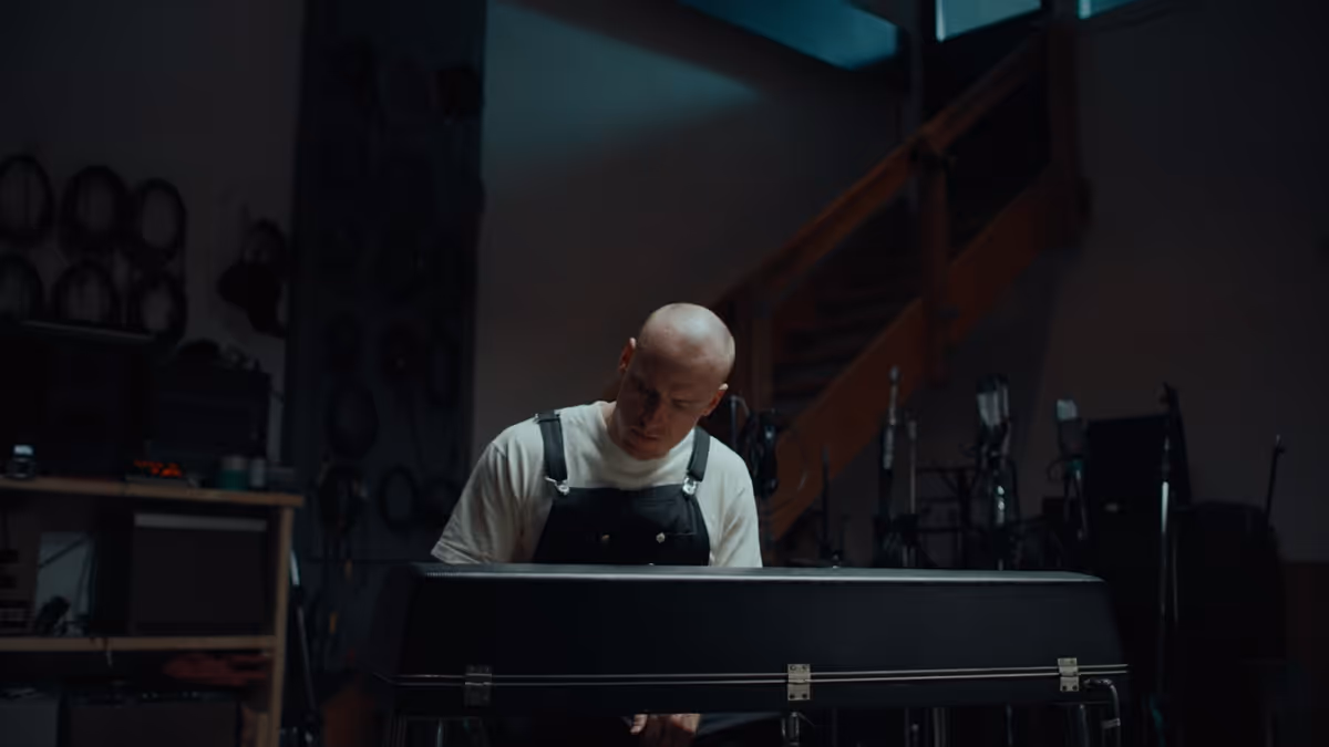 Man in white shirt and black overalls opening a black guitar case in a dimly lit room with equipment and staircase in the background.