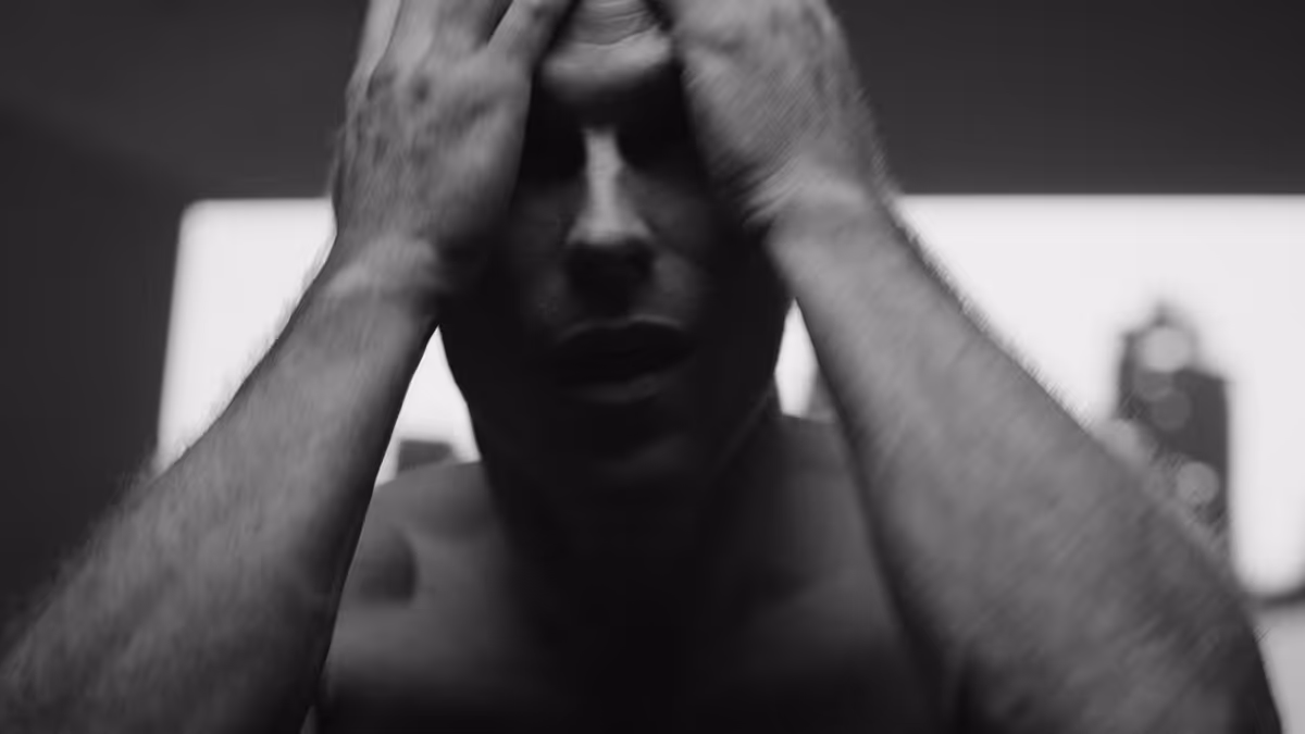 Black and white close-up of a shirtless man holding his head with both hands, expressing distress or frustration.