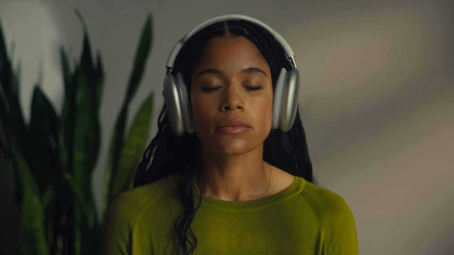 Woman with closed eyes wearing silver over-ear headphones and a green sweater, with a blurred plant in the background.