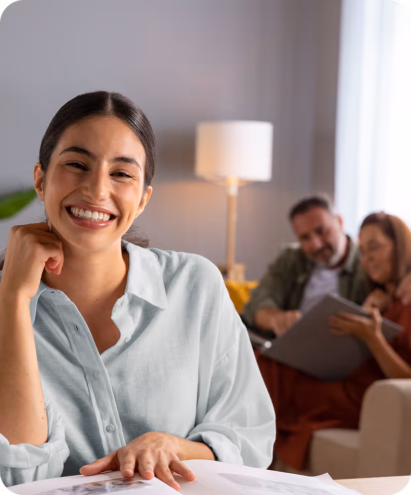 Smiling woman sitting at a table with documents, with two people discussing paperwork in the background.