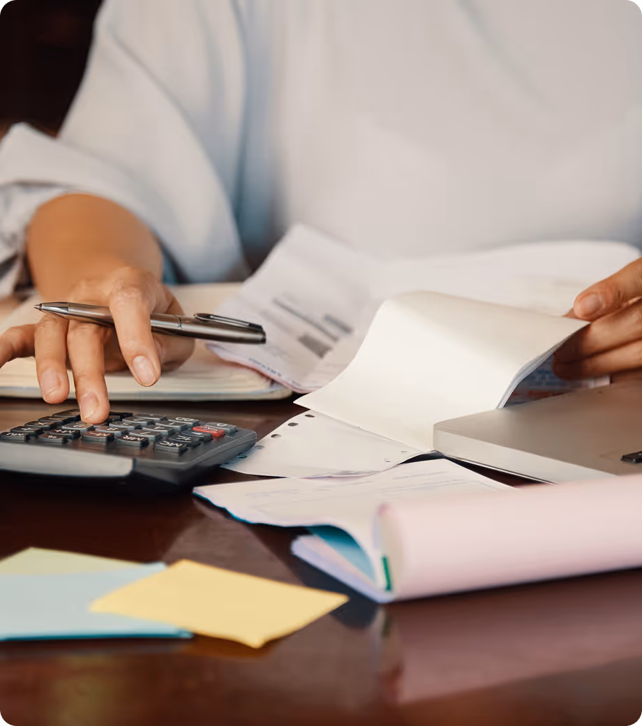 Close-up of hands using a calculator and reviewing financial documents at a desk with papers and a laptop.