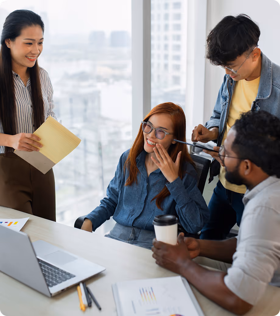 Group of four colleagues gathered around a desk in a bright office, smiling and discussing work together.
