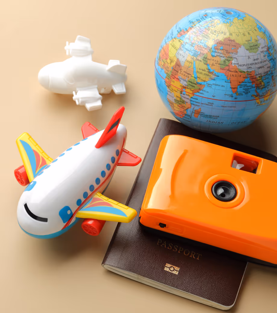 Flat lay of travel-themed objects including a colorful toy airplane, a white plastic airplane, a small globe, a brown passport, and a bright orange toy camera on a beige background.